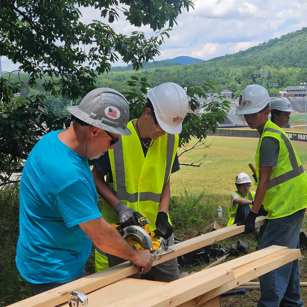 Three carpentry workers in helmets, two with lime green vests and one with turquoise t-shirt, work on sawing boards at a work site, with campus landscape in background