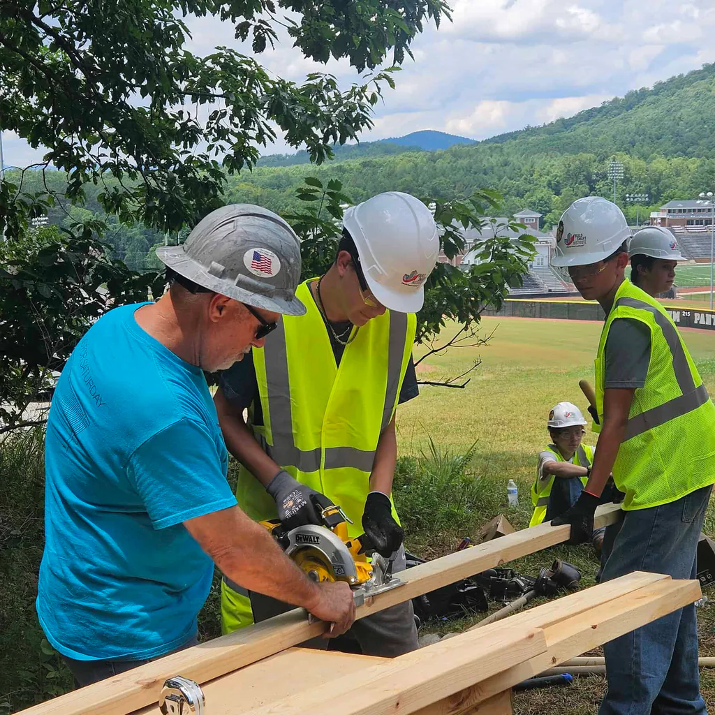 Three carpentry workers in helmets, two with lime green vests and one with turquoise t-shirt, work on sawing boards at a work site, with campus landscape in background
