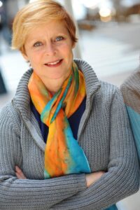 Janine Latus head shot, standing with arms folded, short blond hair, a blue sweater with open collar and scarf with oranges and bright blue