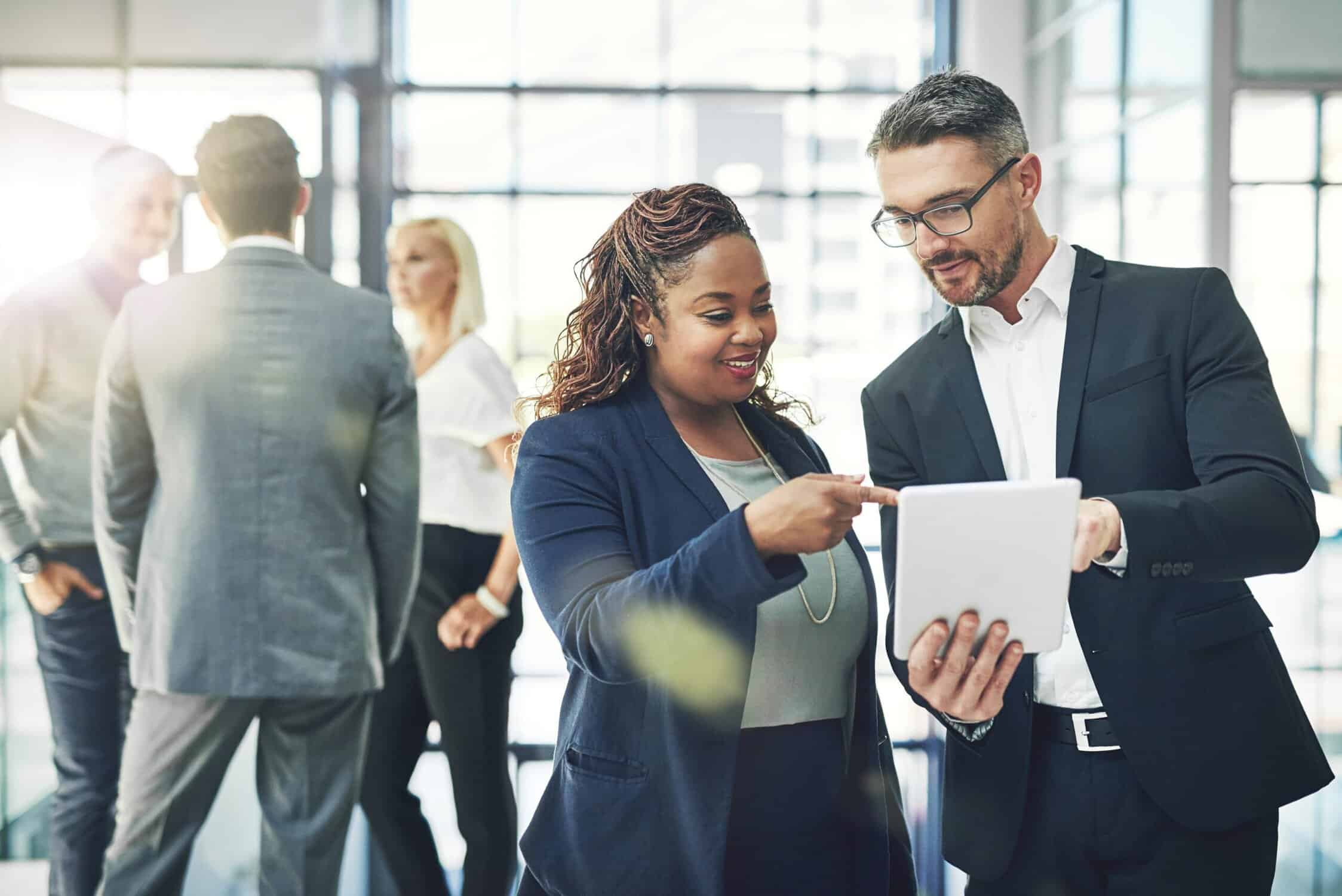 female and male business employees looking over paperwork in hallway while three employees discuss to the side in another group