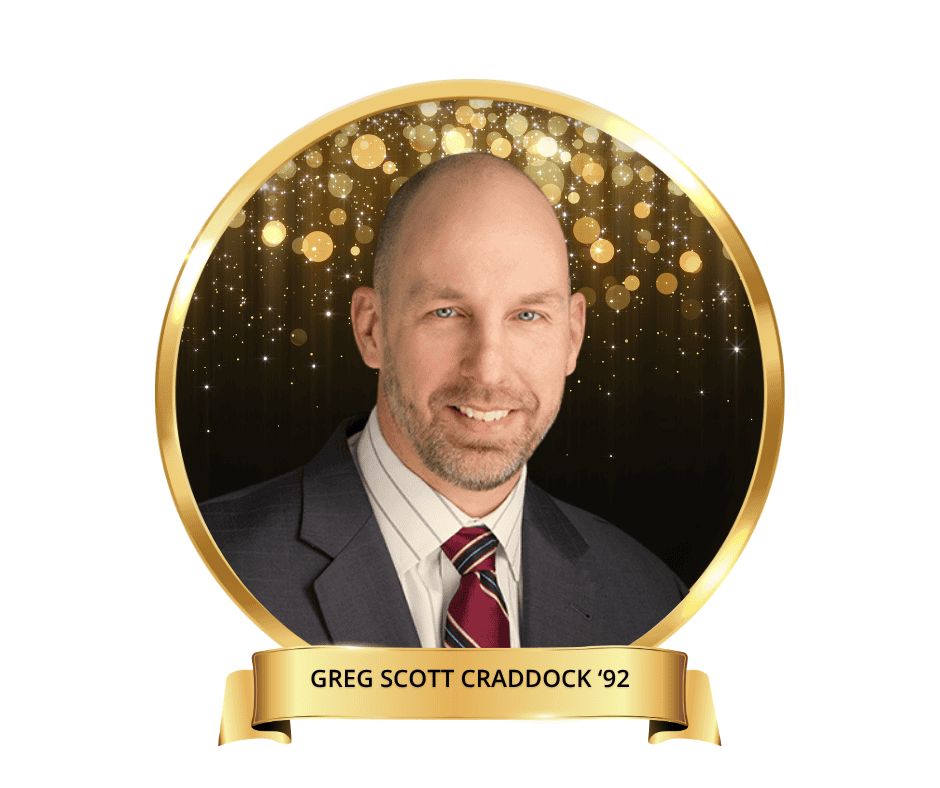 A headshot of Greg Scott Craddock. Wearing a grey suit with a red tie with a black and gold drop.