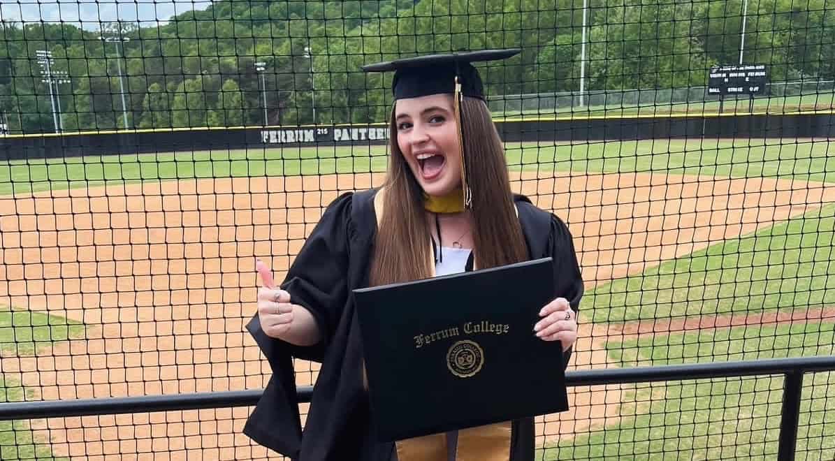 Ashton Lambeth in black cap and gown in front of ball field and fence, holding diploma, with long dark hair and gesturing with thumbs up