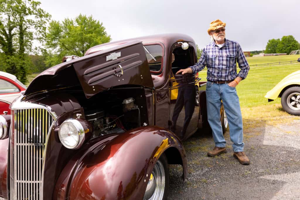 Man in blue jeans, plaid shirt, and light-colored hat stands next to a vintage maroon car with the hood open over the engine, with a bit of BRIM landscape showing in background
