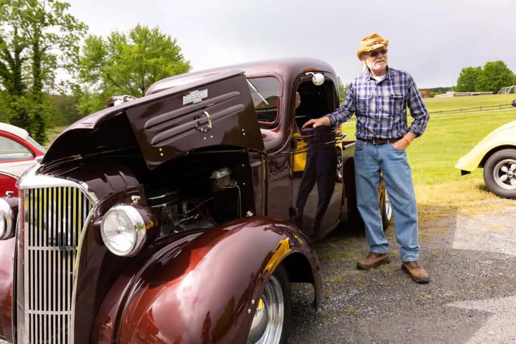 Man in blue jeans, plaid shirt, and light-colored hat stands next to a vintage maroon car with the hood open over the engine, with a bit of BRIM landscape showing in background
