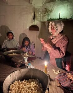 Woman in bonnet and period dress stringing popcorn in 1800 farmhouse with 2 children