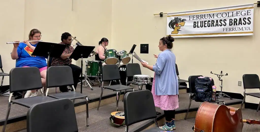 Rehearsal in band room with 3 students with drums, trumpet, and clarinet, with Emily Blankenship-Tucker directing in blue and pink clothes. Sign on wall says Ferrum College Bluegrass Brass.