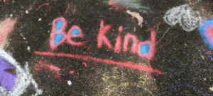 Be Kind written in pink chalk, underlined and with blue dots on a black background, with parts of chalk images such as a butterfly around it.