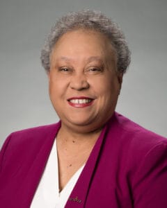 Head shot of Dr. Belle Wheelan on a grey background, with very short hair, a purple jacket, and white blouse.