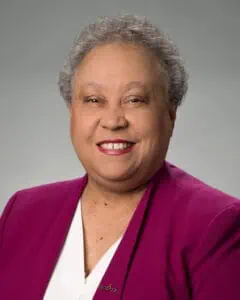 Head shot of Dr. Belle Wheelan on a grey background, with very short hair, a purple jacket, and white blouse.