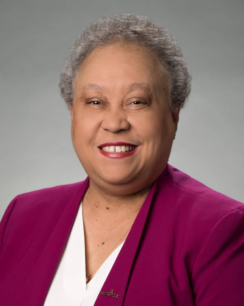 Head shot of Dr. Belle Wheelan on a grey background, with very short hair, a purple jacket, and white blouse.