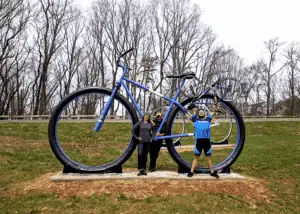 Big Old Bikes sculpture with two giant bikes in park, with Jake Smith and Kathleen in dark clothing, and Tim Pohlad-Thomas in biking clothes and helmet holding his bike up in the air