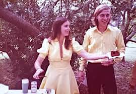 Young Carolyn Thomas and Bob Pohlad sharing wedding cake, both dressed in yellow at their outdoor wedding in 1975 