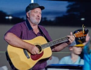 Bobby Radford with guitar in striped shirt and ball cap at BRIM farm pavilion