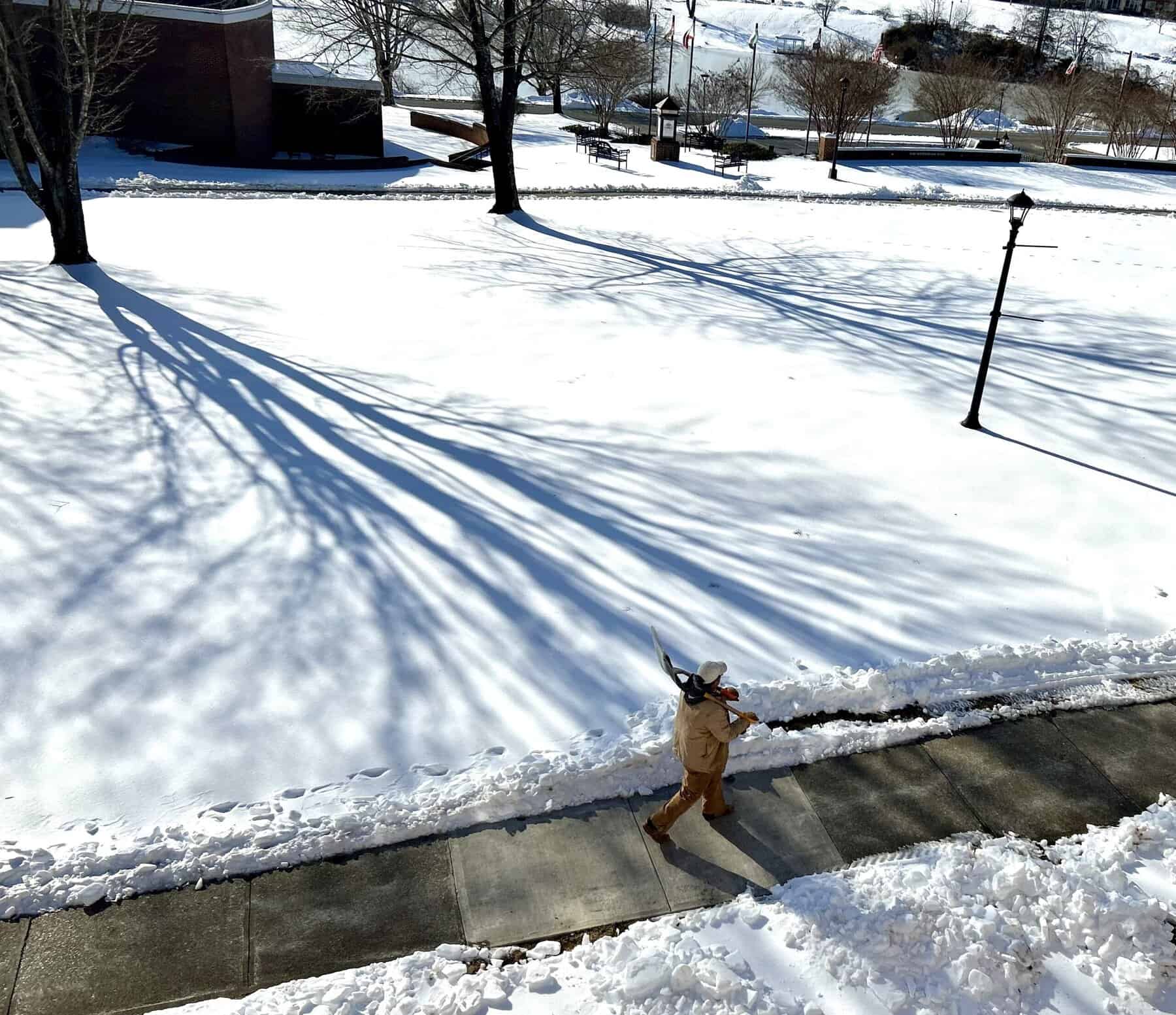 Man in tan winter clothes carrying a shovel down a cleared sidewalk on snow-covered campus with dramatic tree shadows and. lamppost