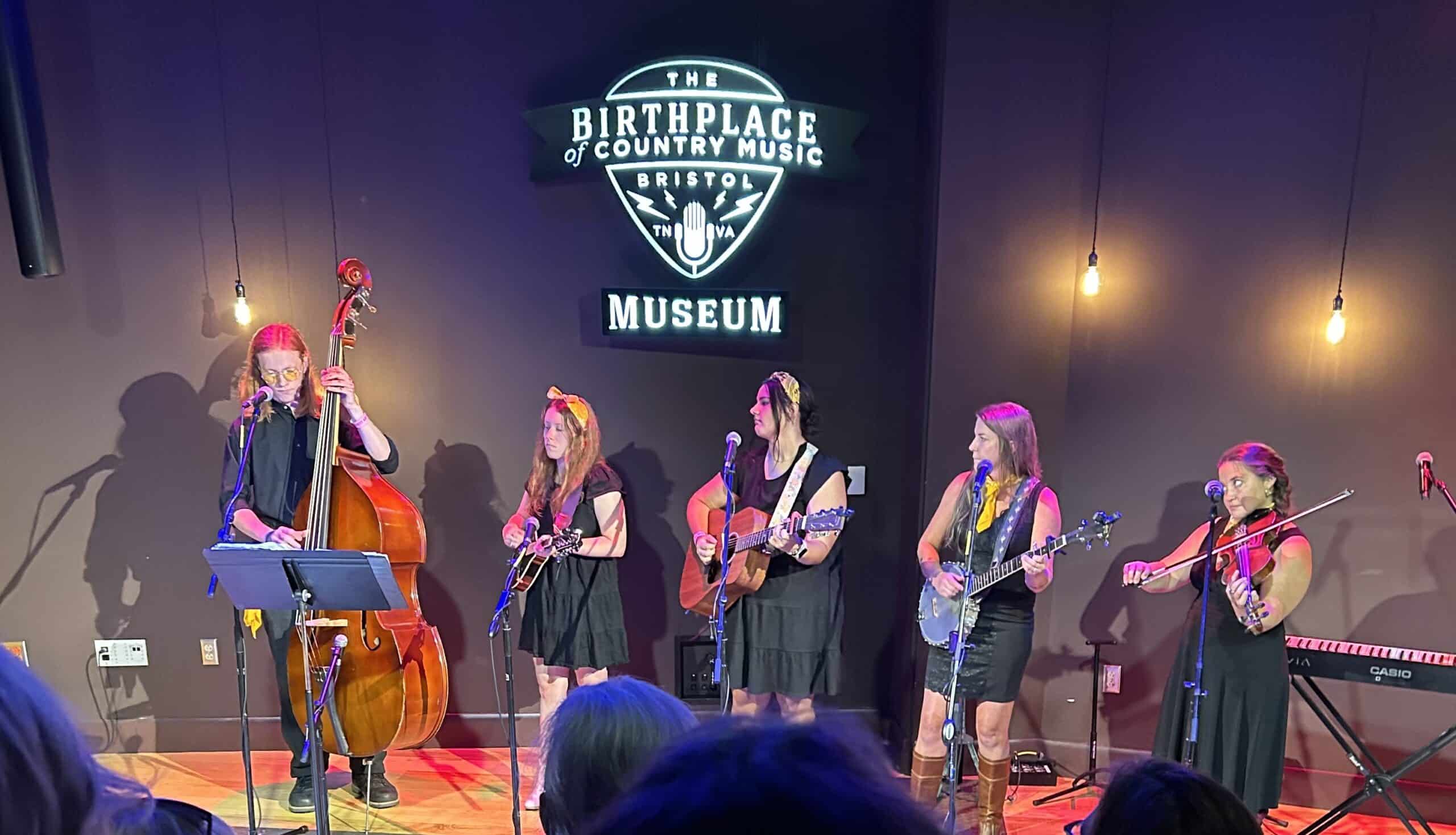 Orchestra Appalachia on small stage, 4 women in black dresses and gold kerchiefs with country instruments, one male on left in black playing base with long red hair. Sign for Birthplace of Country Music in Bristol behind them above.