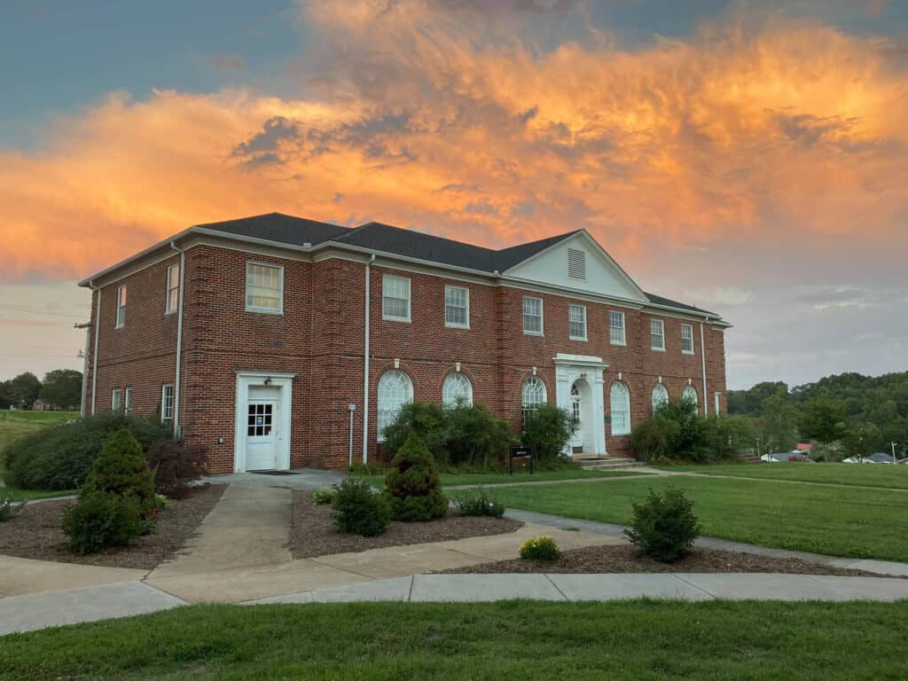 Britt Hall, brick classical building with white trim, 2 stories, with green landscaping and pink sunset clouds above, by Tracy Holley '96
