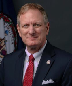 A middle-aged man with short light brown hair wearing a dark blue suit, white shirt, and red patterned tie, with a Virginia state flag in the back.
