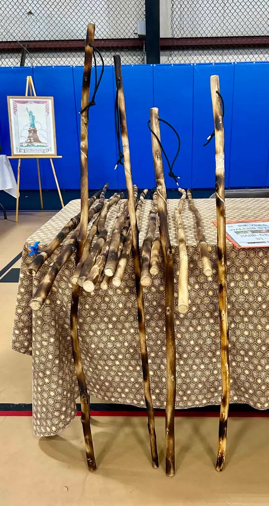 Group of tall walking sticks for sale at festival leaning against a wooden table with blue wall and Statue of Liberty fabric art in background