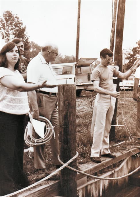 Black and white photo from 1980s of 3 adults standing at a boat dock in summer clothes, one woman with short dark hair on left and two men