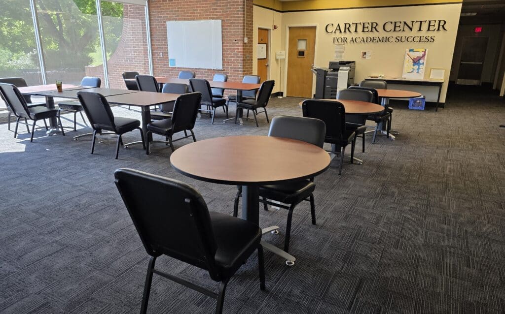 Carter Center main room with round tables and chairs, dark carpet, back brick wall with name of center, windows on left