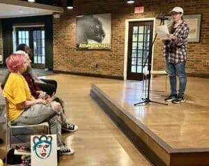Mattie Green, in jeans, plaid shirt, and ball cap, is reading from a paper while standing at a microphone on the low stage in the Panthers Den at Ferrum College. Wood floor, brick wall with panther painting, windows, and the front row of the audience are also visible. A painting of a face on a white background is in the foreground next to the audience.
