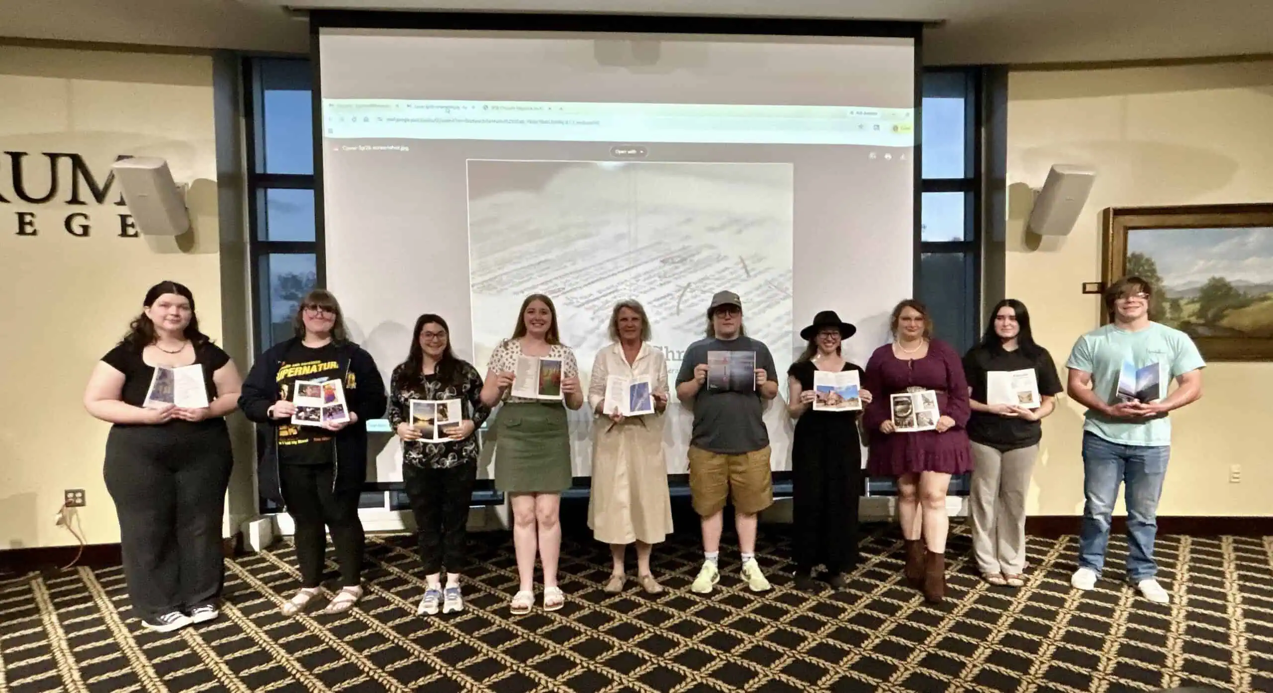 Ten students and one faculty advisor in the middle, holding open copies of a small magazine, are lined up in front of a screen that displays the grey cover of the spring magazine. They stand on a black and gold patterns carpet in a room with gold walls and art on the walls.