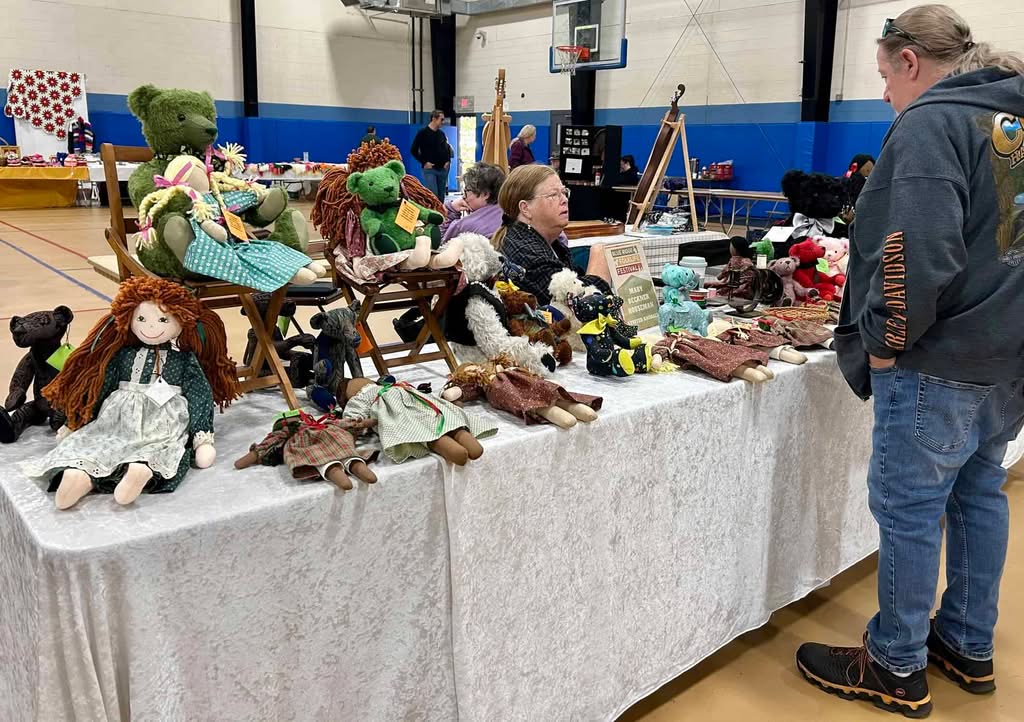 Two women sitting back to back at tables with white tablecloths covered with cloth toys--some large teddy bears and colorful dolls. A man stands on right. Other crafts and shoppers are in back, with gym walls and basketball hoop.