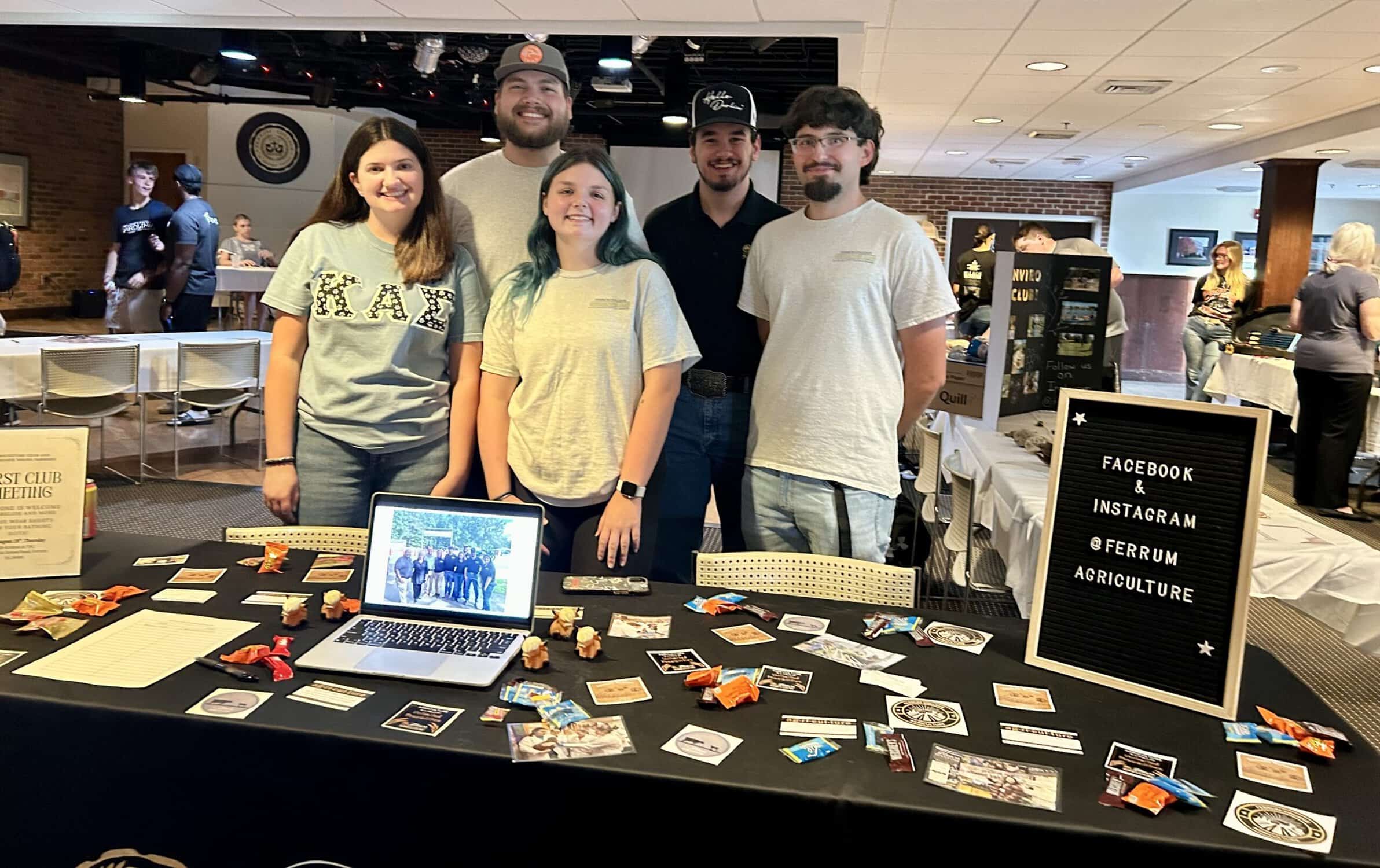 Five students in t-shirts standing behind a table with laptop and lots of stickers and club fair items on black tablecloth, with more students in background