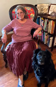 Woman with gray hair in maroon shirt and shirt sitting in vintage wooden chair with red flowered cushions, beside black dog, with full bookcase behind them