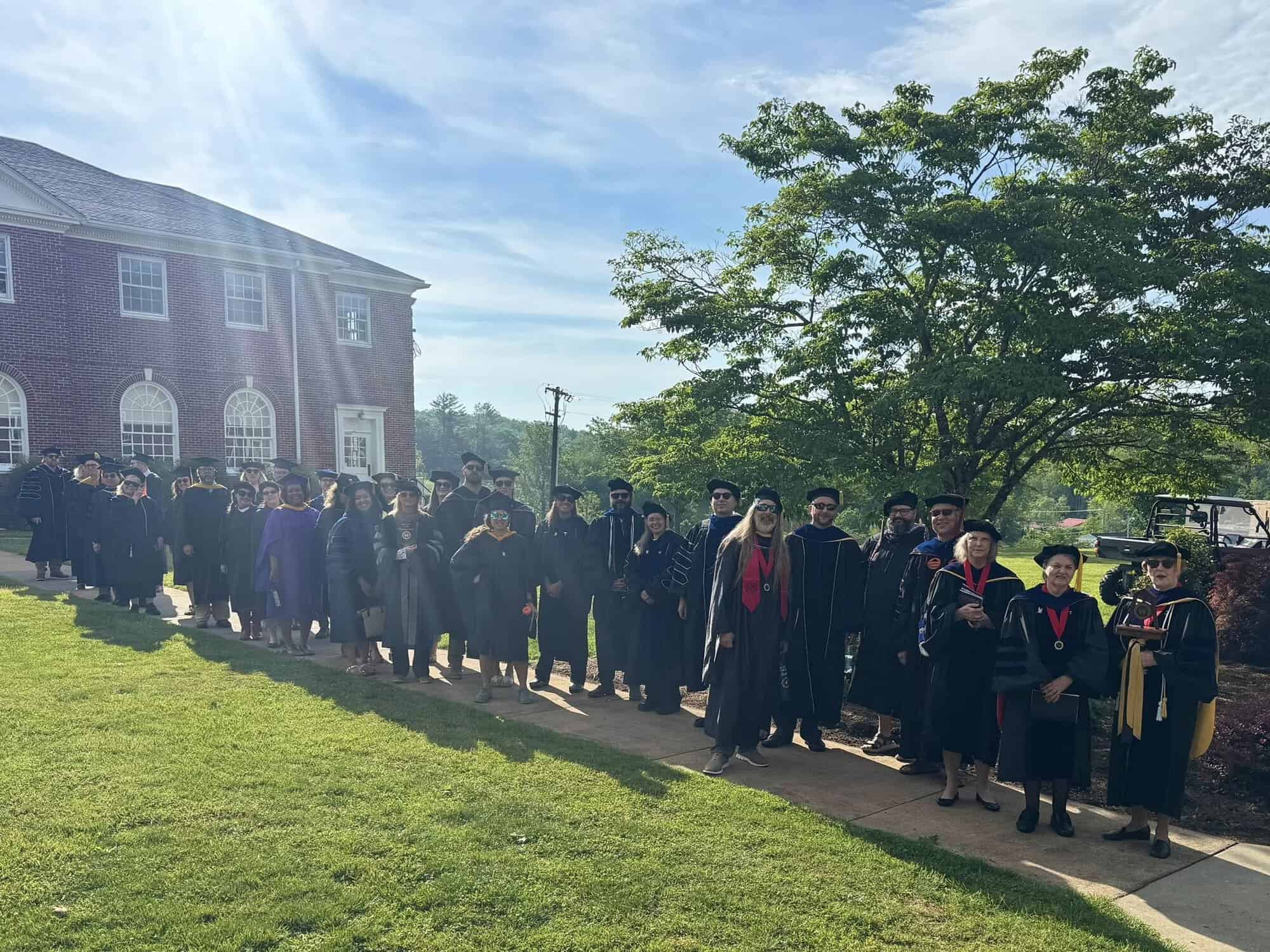 Faculty members in academic regalia linked up on sidewalk in front of brick building with grass, trees, and blue sky around them