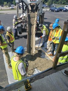 Several men in construction gear with bright blue hard hats and neon lime green vests surround a structure that is pouring concrete in an outdoor space
