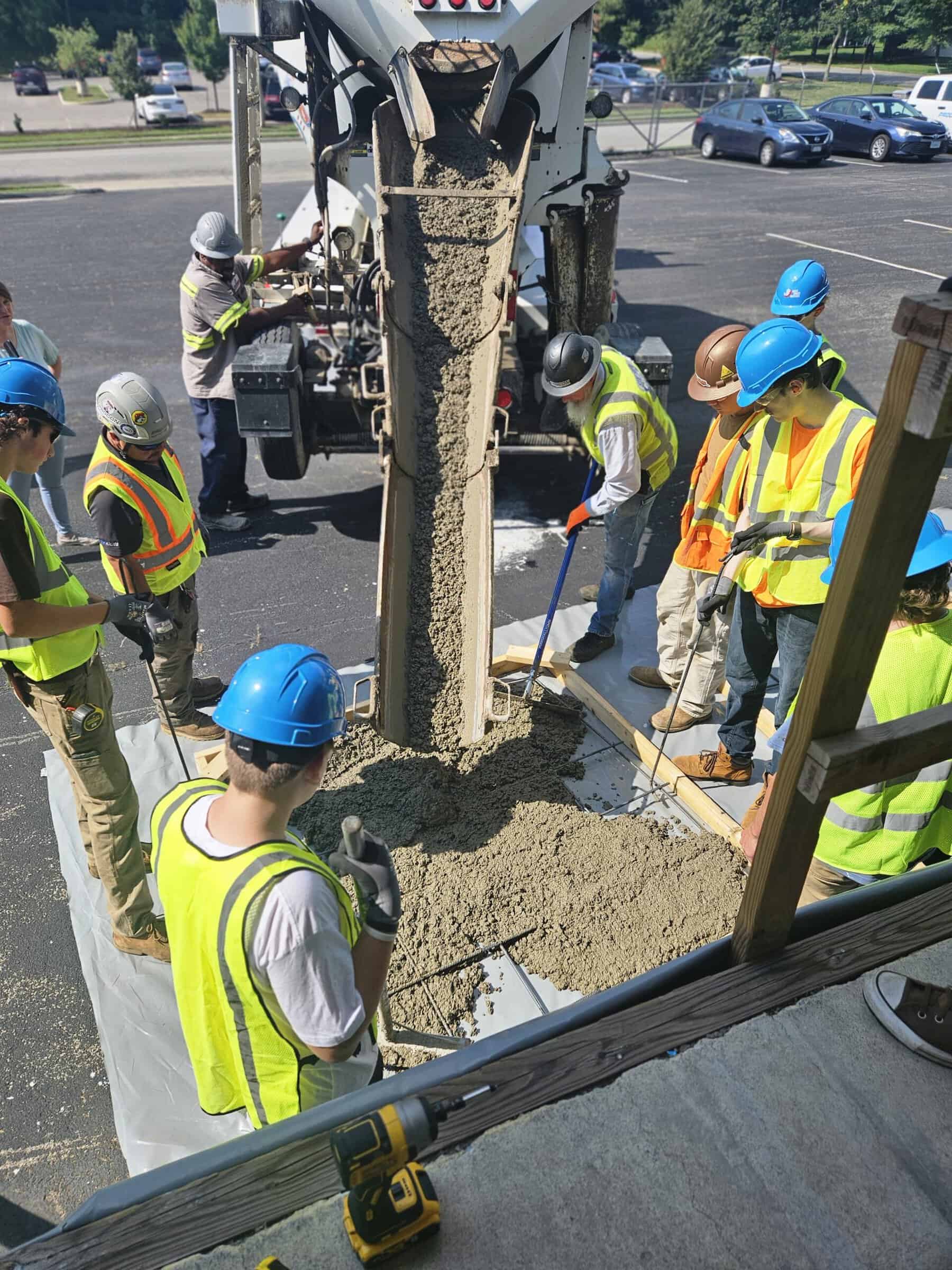 A group of people in construction gear with bright blue hard hats and neon lime green vests surround a structure that is pouring concrete into a mold in an outdoor space