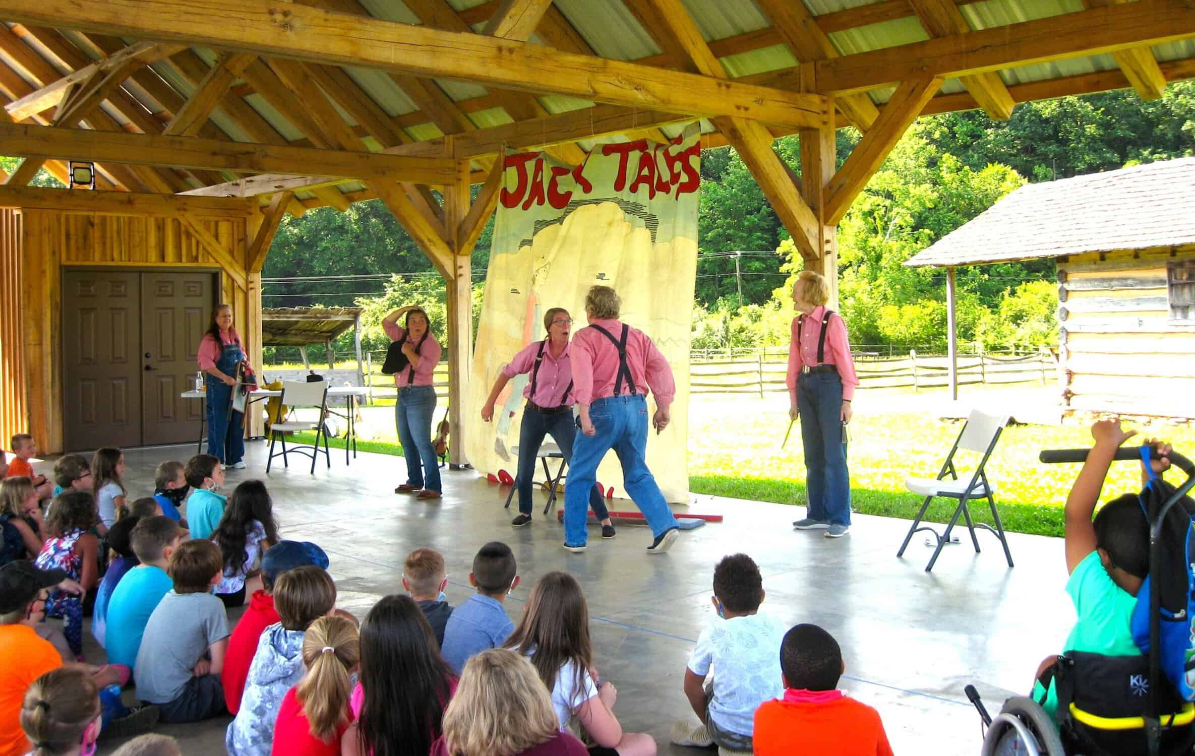 Five performers in red checked shirts and jeans dramatize a folktale with Jack Tales banner in farm museum pavilion for small school children