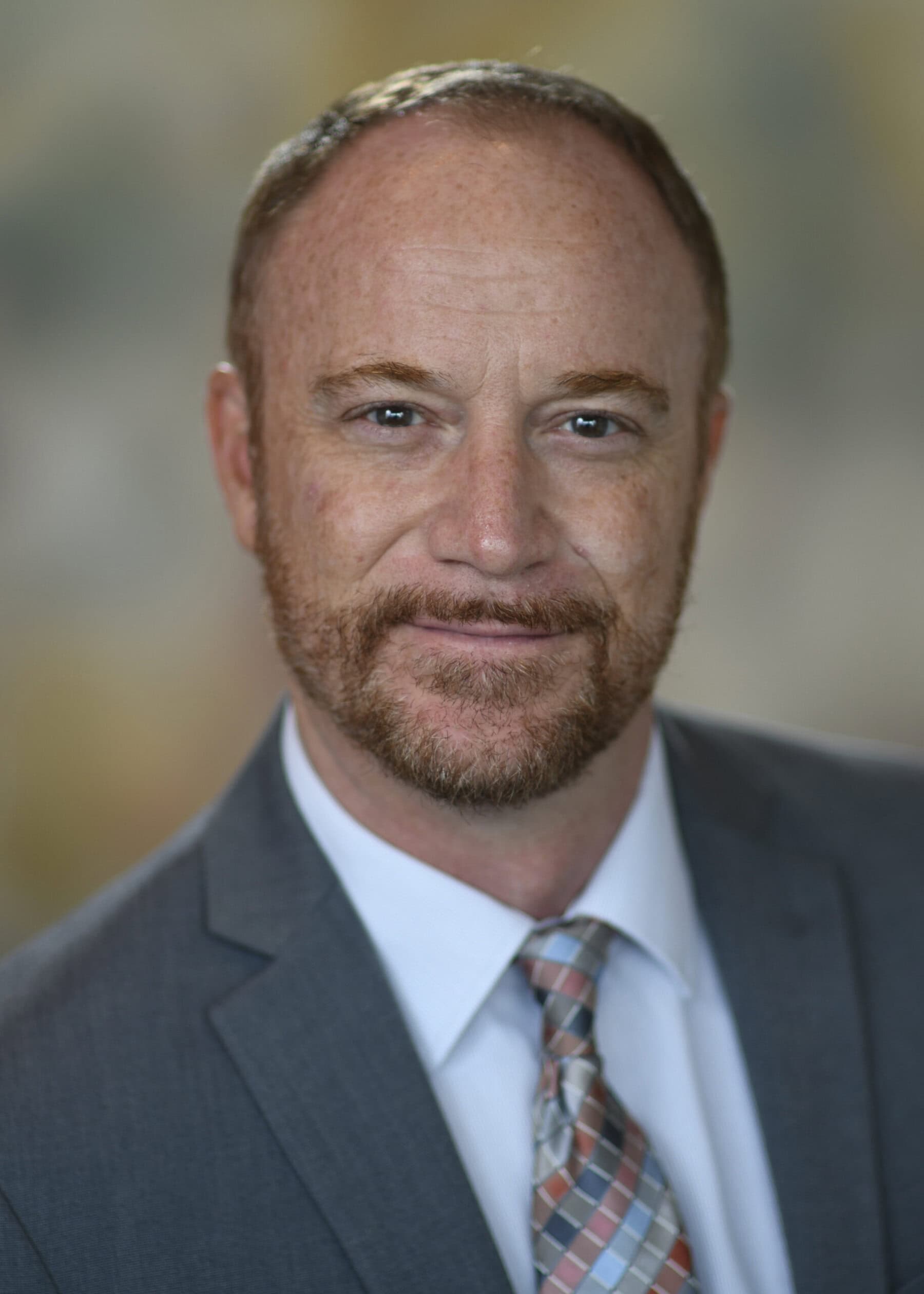 Head shot of Chris Whitlow with dark jacket, white shirt, tie with several colors, some facial hair and dark hair, with grey background