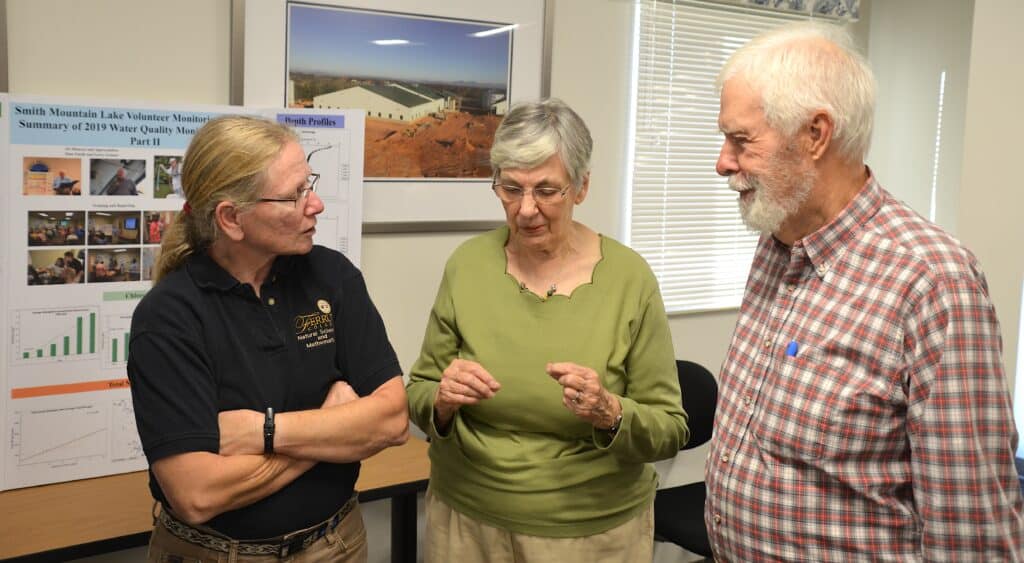 Carol Love in blonde hair and navy shirt talking to two older adults on her right, with poster and painting behind them in a meeting room