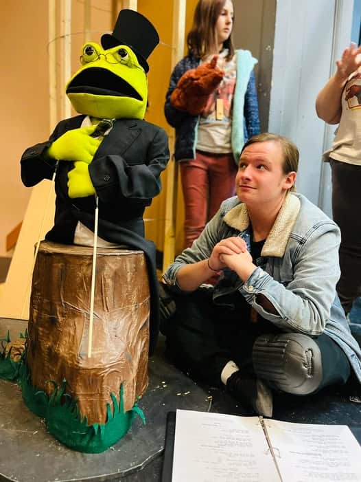 Lime green Doc Bullfrog puppet in a black top hat and jacket sitting on a stump next to Gage Shelton sitting on the stage in light jacket, with other puppeteers partially seen in the background