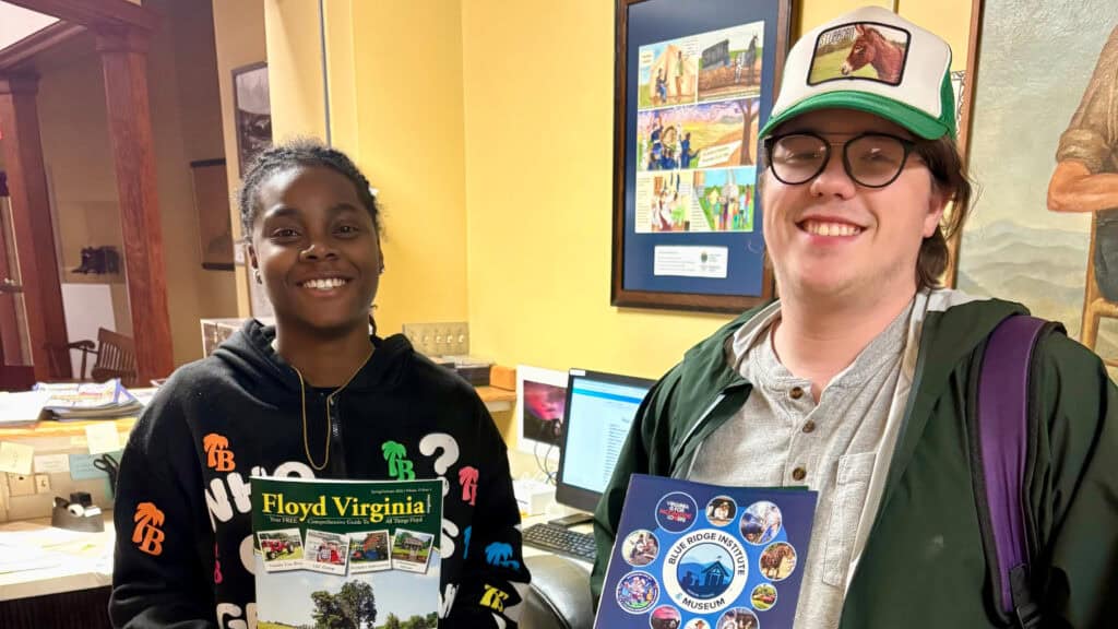 Two students smiling at the camera both holding a book that are about Floyd Virginia and the Blue Ridge Institute & Museum. Both are standing in front of a desk. The wall behind them is yellow and there are paintings on them.