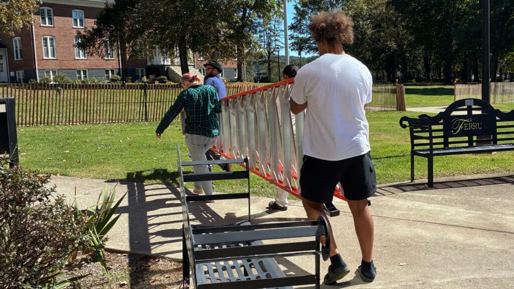 Three students carrying a ladder outside on a sunny day. There are benches to the left and right of them. In front of them is a fence with a opening to the left. The grass is green. The young man in the back is wearing a white shirt with black shorts. The woman holding the front is in a green plaid jacket wearing jeans.  