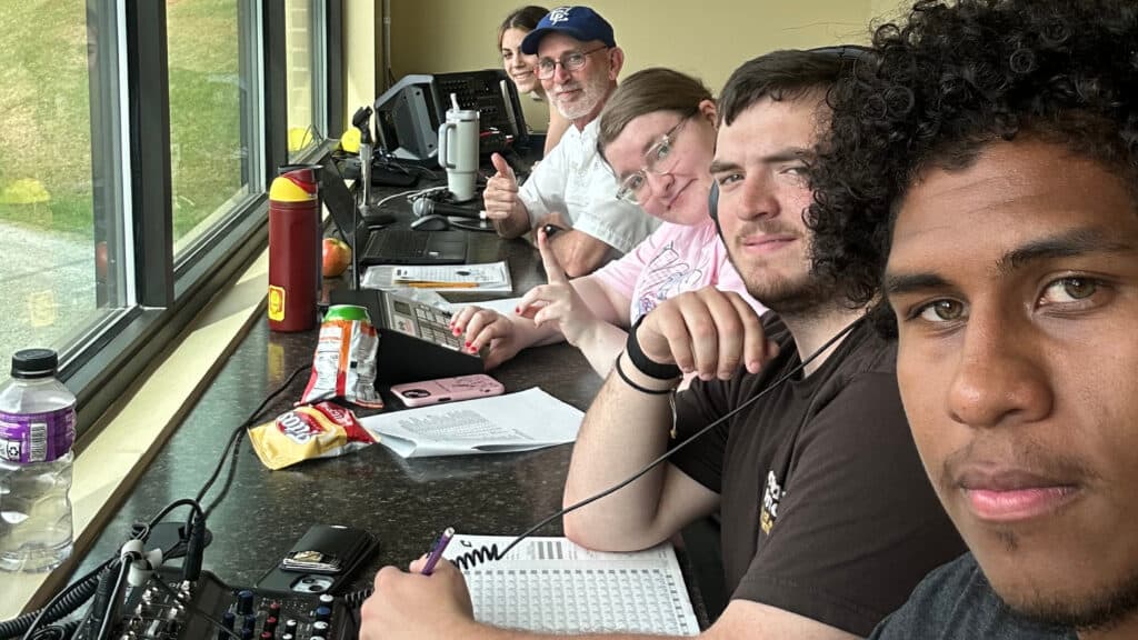 A group of students posing while one student is working the walkup music, the older man is tracking live stats, the young woman next time is updating the scoreboard, next to her the young man is writing down the stats, the young man next to him is broadcasting the game live.