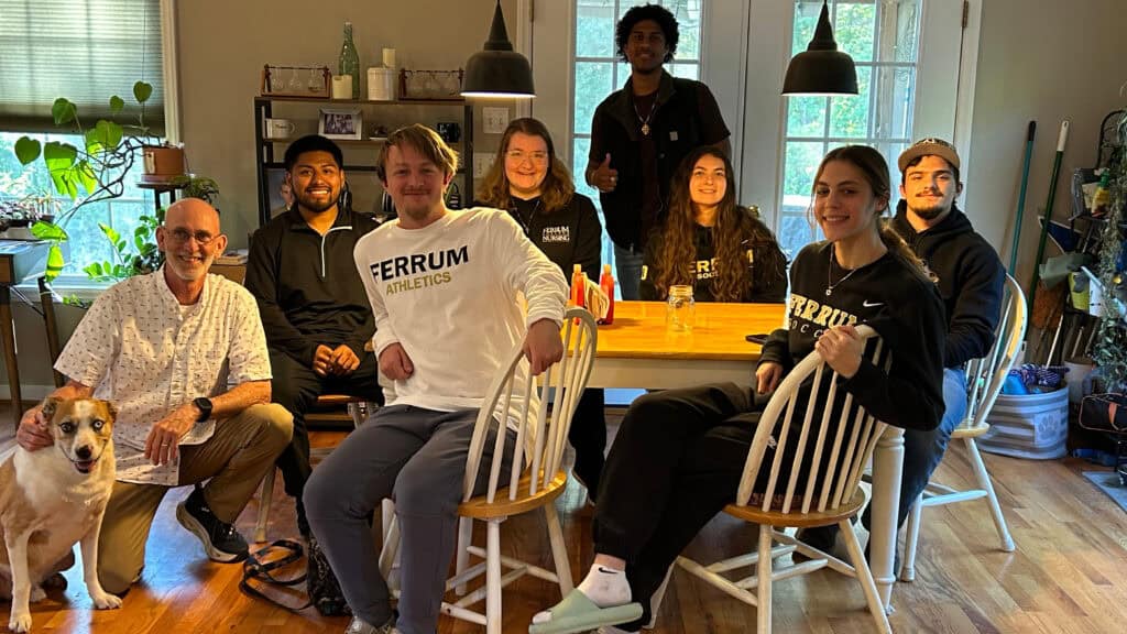 A group of student workers smiling while sitting at a dinner table taking a group photo. There is a dog in the bottom left sitting in front of a man.