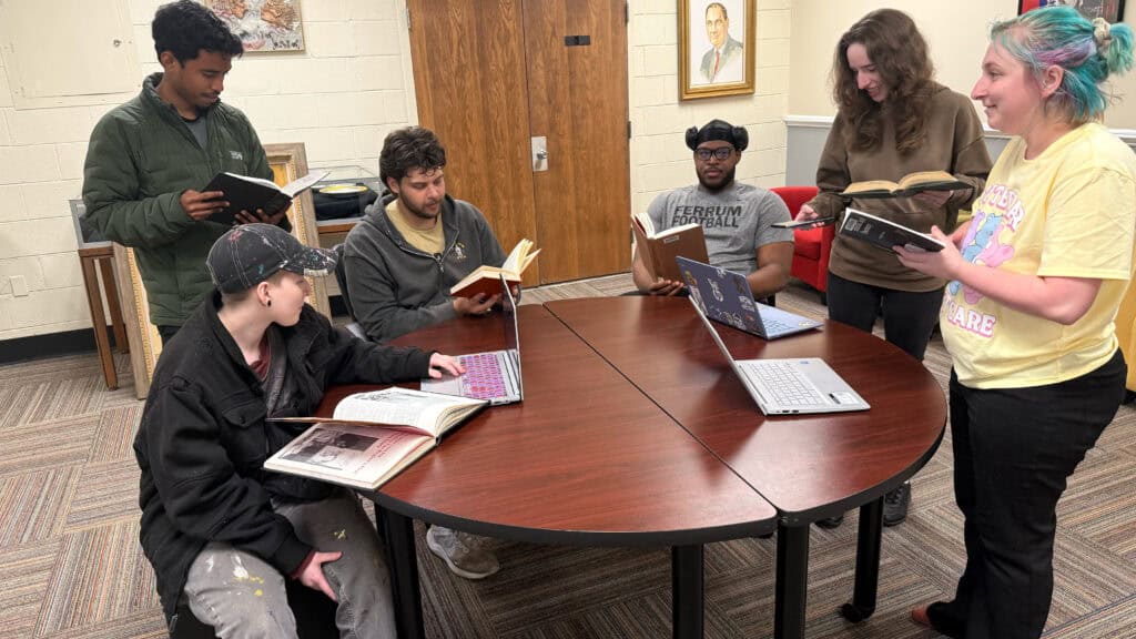 A group of six students standing and sitting around a circular table. They all. are holding a book. There are three laptops on the table. There is a door behind them with a painting on the right and the left with a display case to the left.