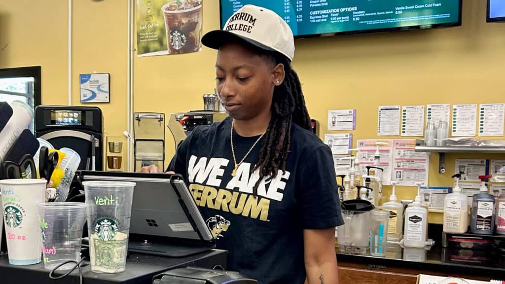 A young woman working behind the cash register. there is a tv screen showing all the different prices and below it is all the different bottles of flavors. On top of the cash register is the different sizes of cups available for purchase.