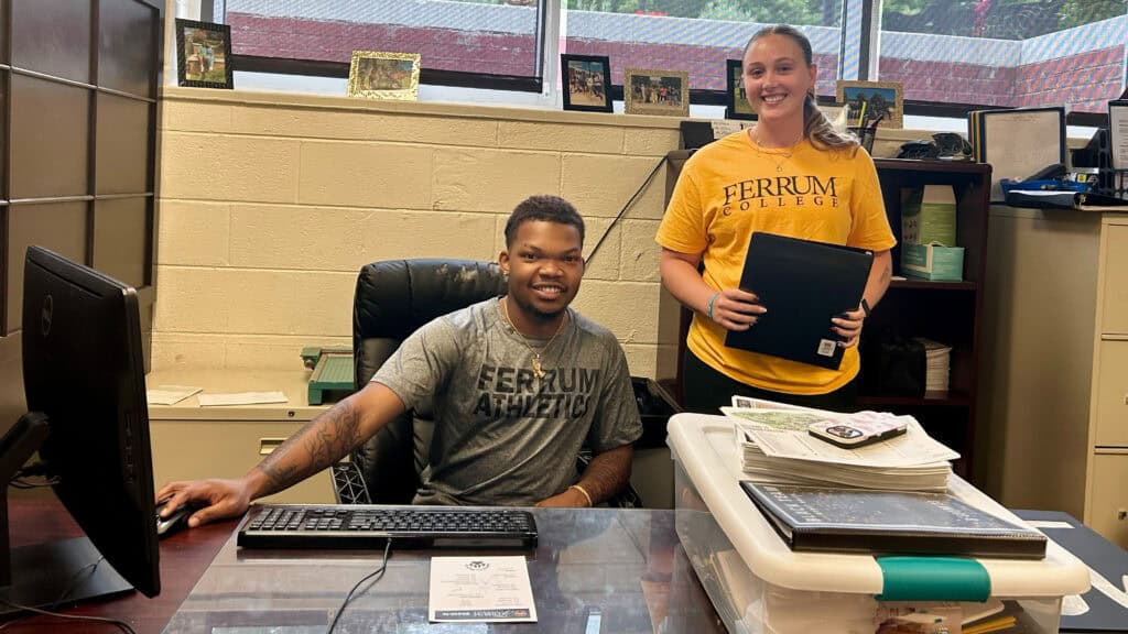 Two student workers smiling at the camera. The young man is sitting down at the desk with his hand on the mouse wearing a gray shirt with Ferrum Athletics across the chest. The young man standing next to him is wearing a dark yellow shirt with Ferrum College across the chest. She's holding a yellow binder. 