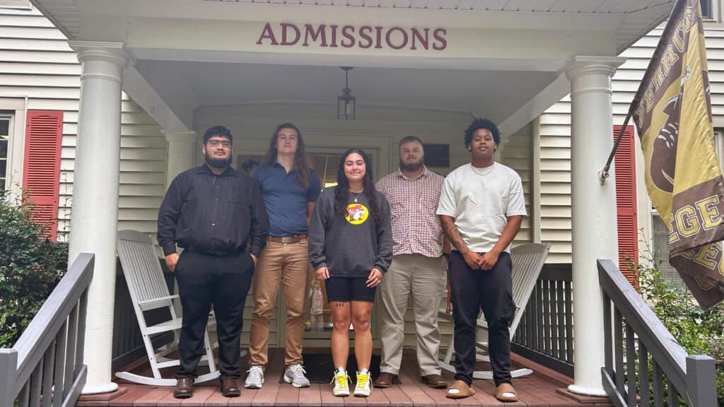 Five student workers posing for a photo. They are standing on the porch of admissions. From left to right: He's wearing a black button shirt with black khakis, the young man next to him he's wearing a blue collard shirt wearing brown khakis, the young woman in the middle is wearing a black sweatshirt with a beaver in the middle wearing black spandex shorts, next to her on the right is a young man wearing a shirt with red lined with white squares in between the lines, on the right next to him is a young man wearing a white colored shirt wearing black pants.