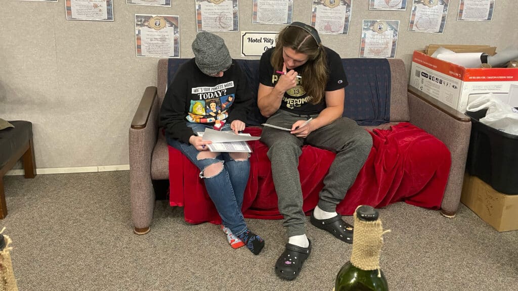Two students sitting on a couch both looking at a paper. They both have their own pieces of paper. There are pieces of paper behind them hung up on a gray wall. On the right of the man is a box filled with paper and paper rolls. 