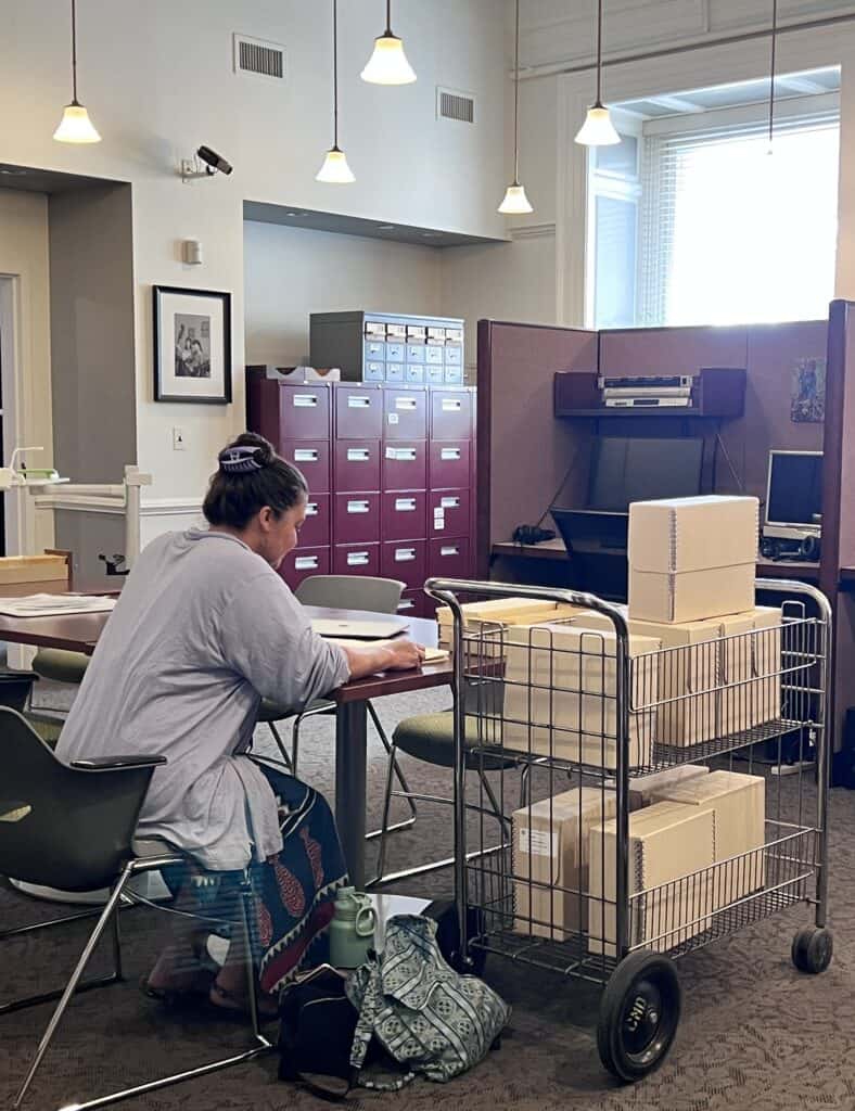Woman in long light blue shirt and dark blue patterned dress show from side and back sitting at a table in an archive, next to a cart full of tan archive boxes