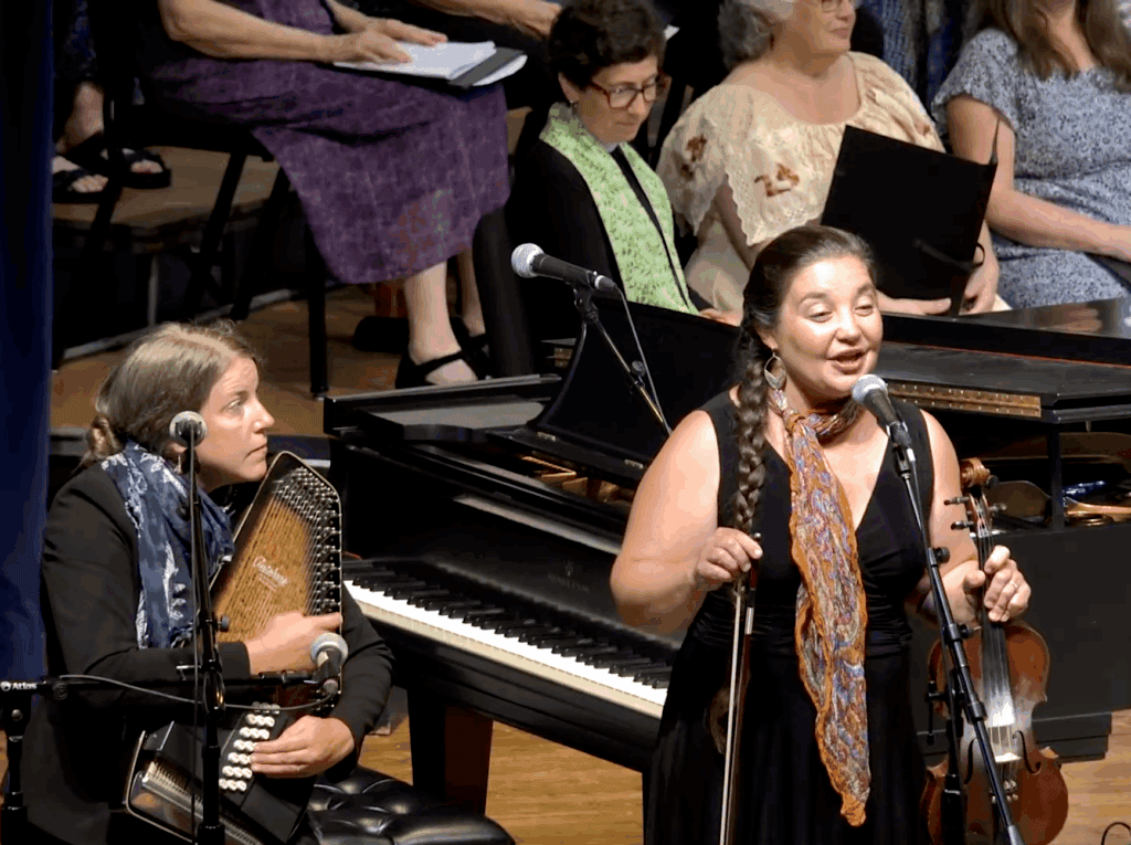 Two women in black clothes, colored scarves, and long hair, one playing autoharp on left, seated, one singing and holding fiddle on right, standing, with piano and seated participants behind them, including one in black with light green priest's stole.