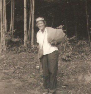 Black and white photo of Emma Gatewood with tree trunks behind her, in dark pants and white shirt, carrying cloth bags