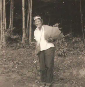 Black and white photo of Emma Gatewood with tree trunks behind her, in dark pants and white shirt, carrying cloth bags