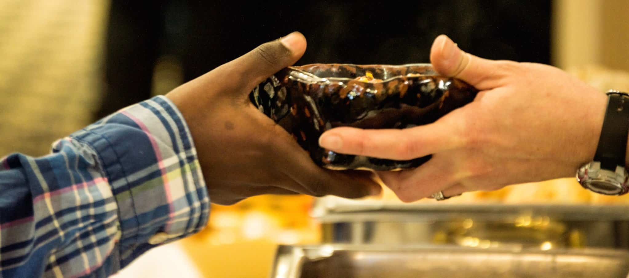 Two hands holding an empty clay bowl on opposite sides of the bowl, with a plaid cuff on one and a watch on the other wrist, and a muted background